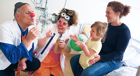 Zwei Clowns machen eine Seifenblase und ein Mädchen mit ihrer Mutter sitzen auf einem Krankenbett und schauen dabei zu