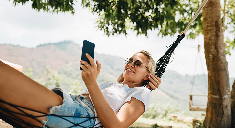 Blonde Frau mit Sonnenbrille liegt draußen in einer Hängematte und schaut in ihr Smartphone