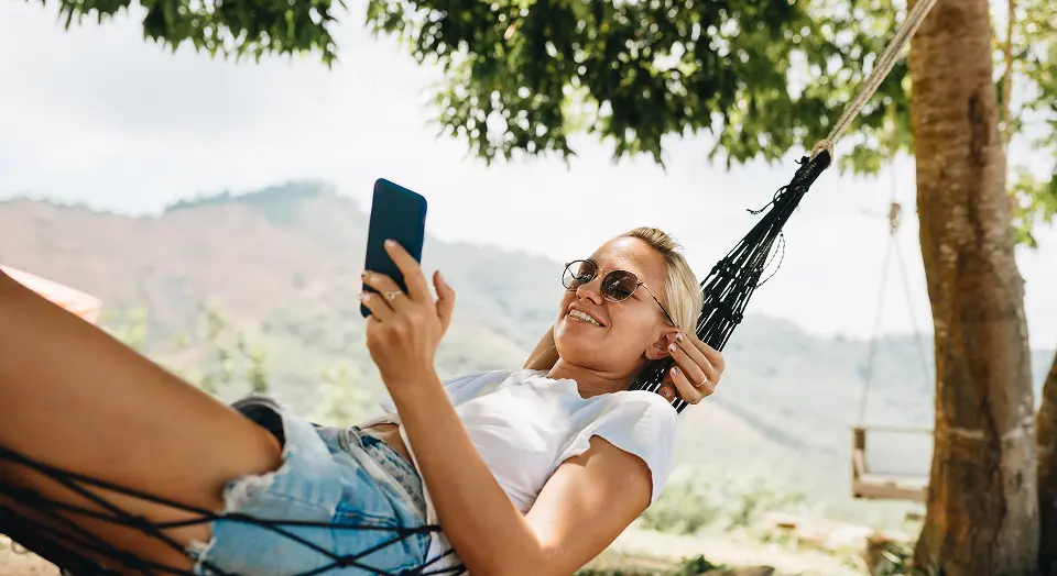 Blonde Frau mit Sonnenbrille liegt draußen in einer Hängematte und schaut in ihr Smartphone
