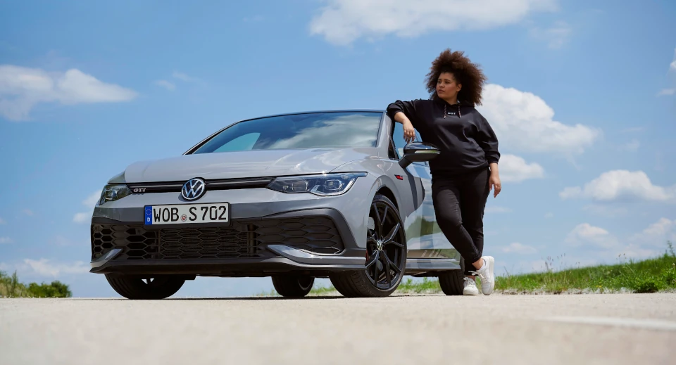 Frau mit Afro steht neben einem neuen VW Auto und lehnt lässig an der Tür