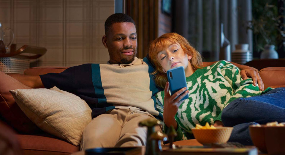 Zwei Personen sitzend aneinander gelehnt auf dem Sofa und schauen in ein blaues Smartphone