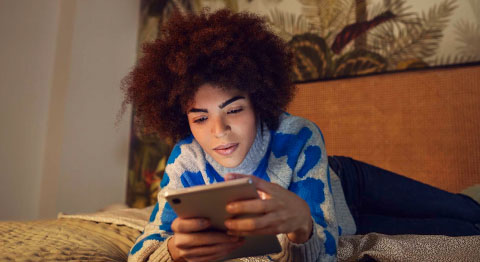 Frau mit blauem Pullover liegt  auf dem Bauch auf dem Bett und blickt auf ihr Tablet