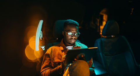 Das Bild zeigt eine Person, die in einem dunklen Auto sitzt und ein Tablet benutzt. Die Beleuchtung ist warm und stimmungsvoll, im Hintergrund sind unscharfe Lichtpunkte zu sehen