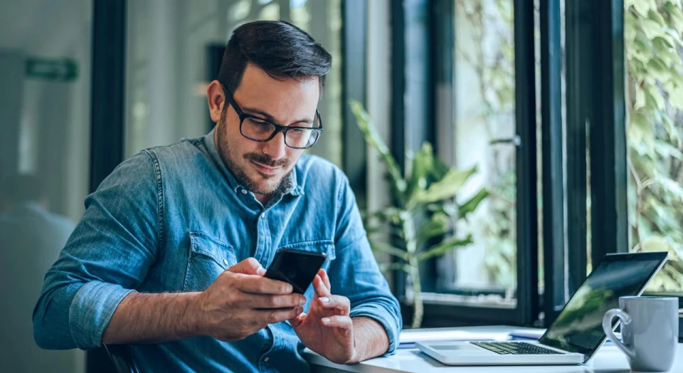 Dunkelhaariger Mann mit schwarzer Brille in Jeanshemd sitzt neben seinem Laptop am Tisch und schaut in sein Smartphone