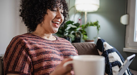 Frau mit schwarzen Locken hält Tasse in der Hand und lacht in ihr Tablet