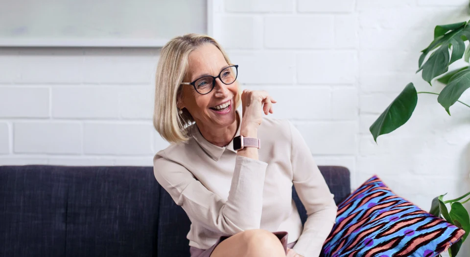Blonde Frau mit schwarzer Brille sitzt auf dem Sofa und lacht