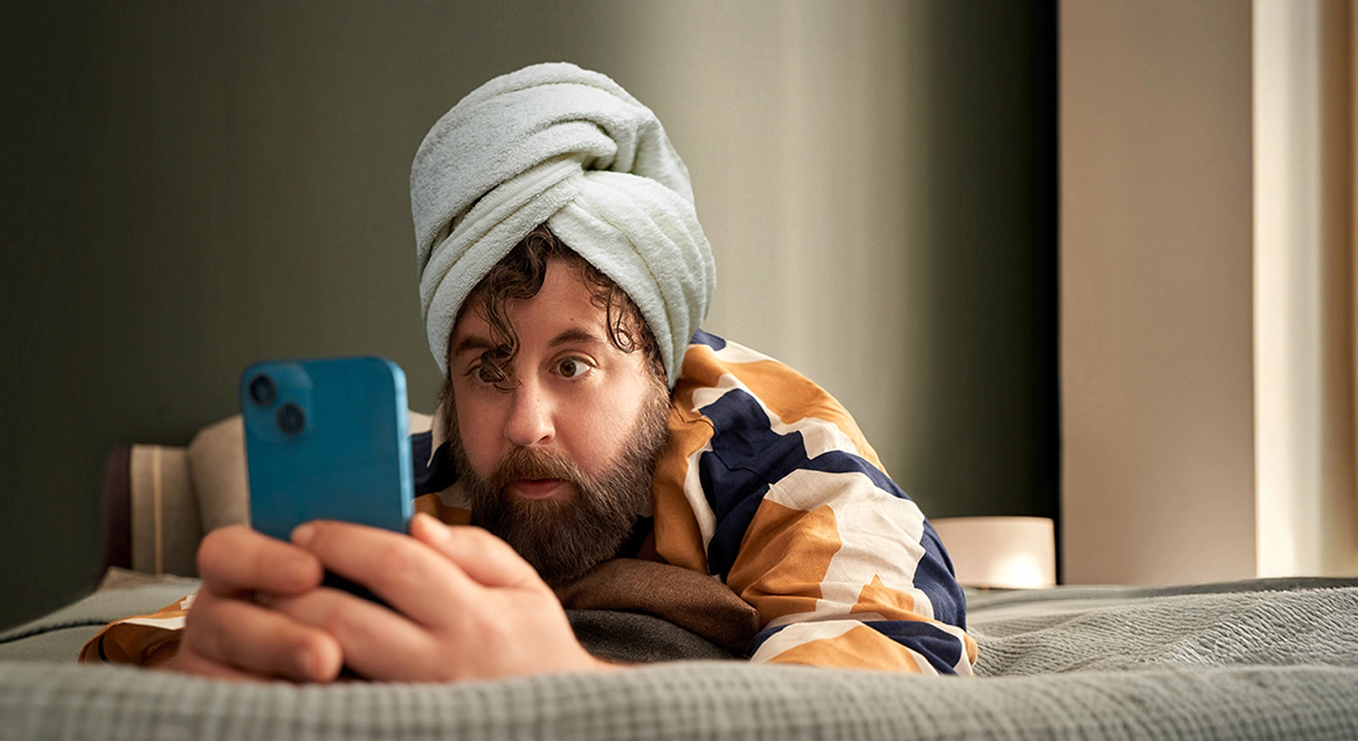 Mann mit Handtuch auf dem Kopf liegt auf dem Bauch auf seinem Bett und schaut in sein Smartphone