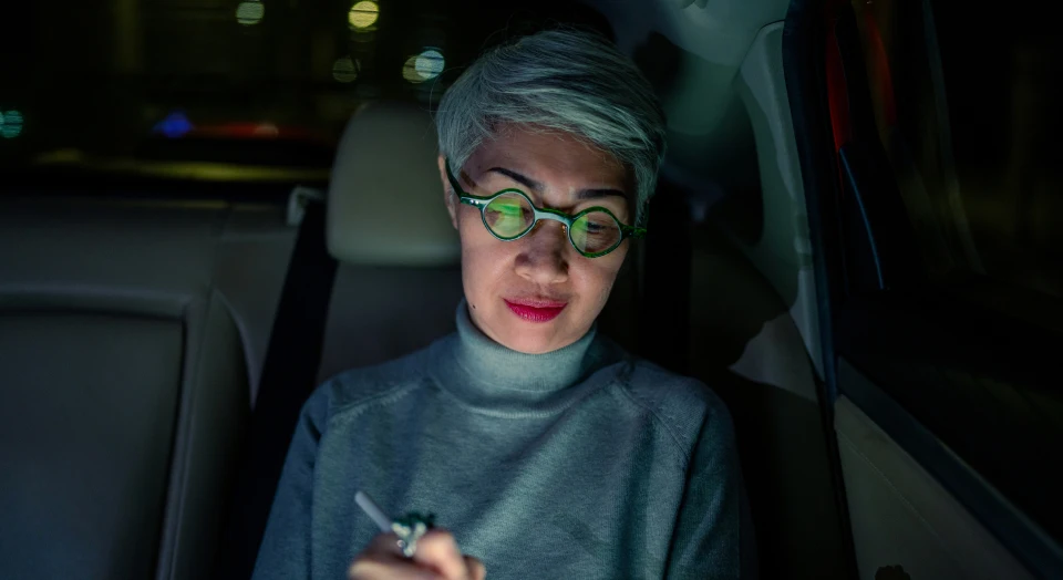 Grauhaarige Frau mit runder Brille sitzt hinten in einem Auto bei Nacht und arbeitet an ihrem Tablet, das sich in ihren Brillengläsern spiegelt