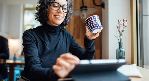 Lockige Frau mit schwarzer Brille hält Tasse in der Hand und schaut in ihr Tablet