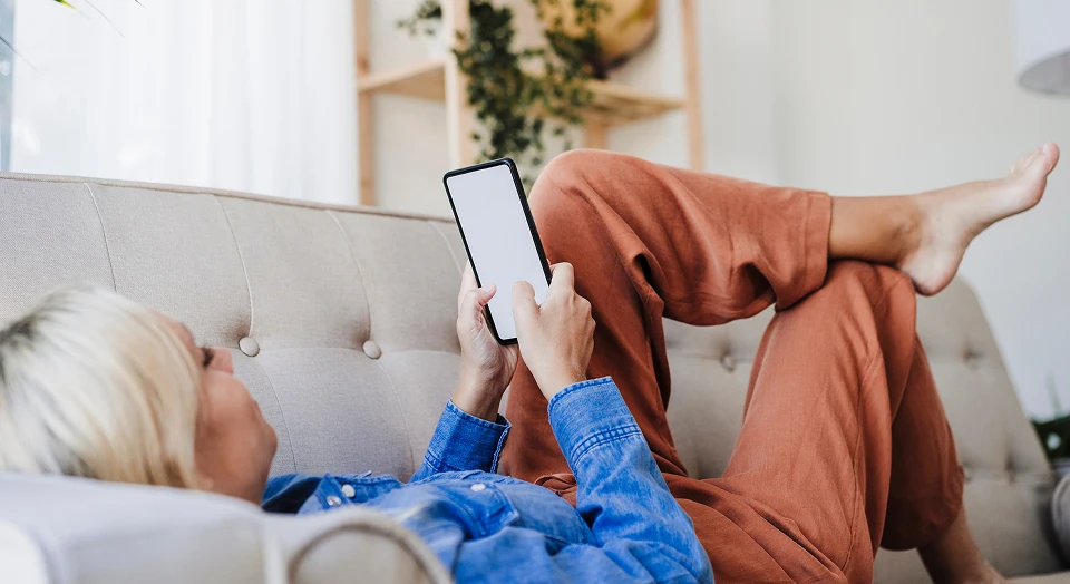 Blonde Frau in Jeansjacke liegt mit überschlagenen Beinen auf dem Sofa und schaut in ihr Smartphone