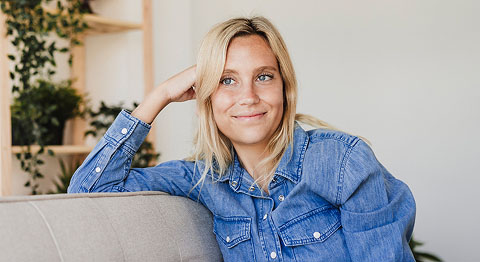 Blonde Frau in Jeansjacke stützt ihren Kopf auf ihrem Arm ab und schaut lächelnd in die Ferne