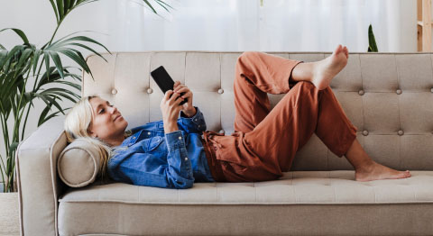Blonde Frau in Jeansjacke liegt auf einem beigen Sofa und schaut in ihr Smartphone