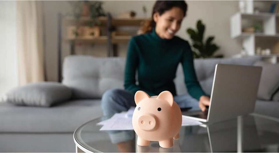 Sparschwein steht auf dem Tisch und im Hintergrund sitzt eine Frau in grünem Oberteil auf dem Sofa und arbeitet an ihrem Laptop