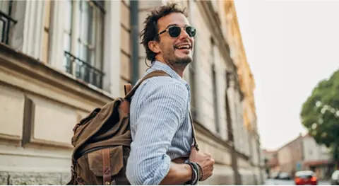 Mann mit Sonnenbrille in Jeanshemd mit Rucksack auf dem Rücken läuft draußen herum und schaut nach hinten