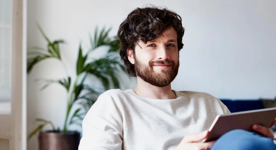 Dunkelhaariger Mann mit Locken in weißem Oberteil sitzt auf einem Sofa und hält ein Tablet in der Hand