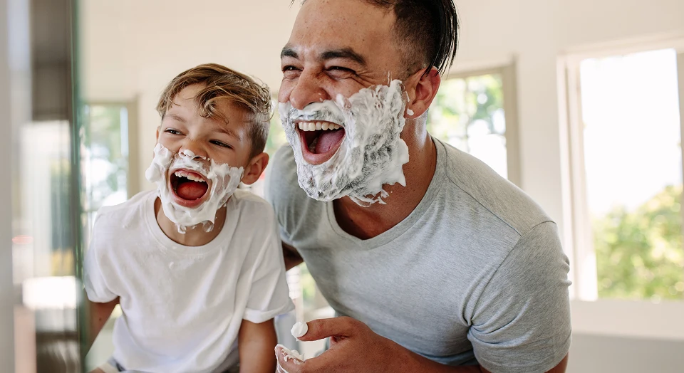 Vater und Sohn stehen lachend mit Rasierschaum im Gesicht im Badezimmer