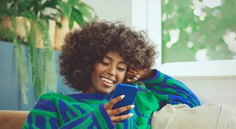 Lockige Frau in buntem Pullover hat blaues Smartphone in der Hand