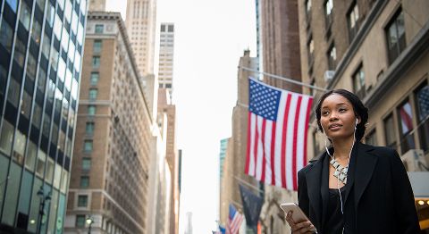 Dunkelhaarige Frau läuft in einer Stadt und hinter ihr weht eine USA-Flagge