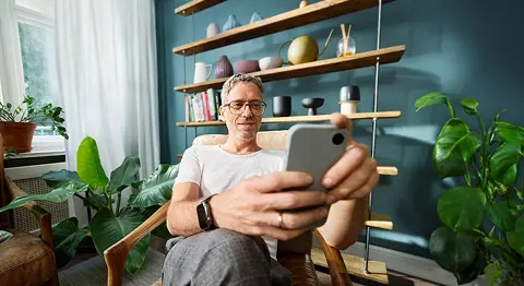Grauhaariger Mann mit Brille sitzt auf einem Sessel und schaut in sein Smartphone