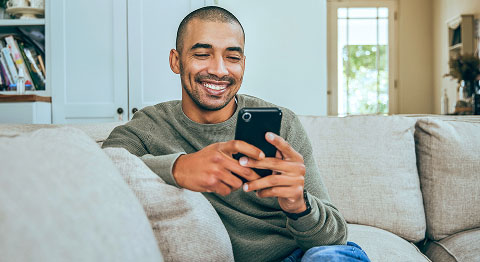Mann in Pullover sitzt auf beigem Sofa und lächelt in sein Smartphone