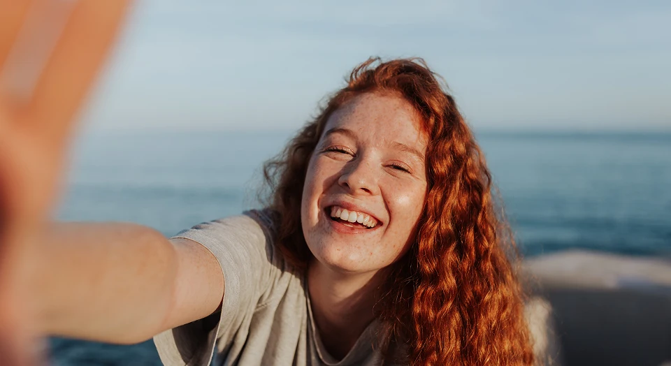 Teenager mit lockigen Haare lacht am Strand in die Kamera