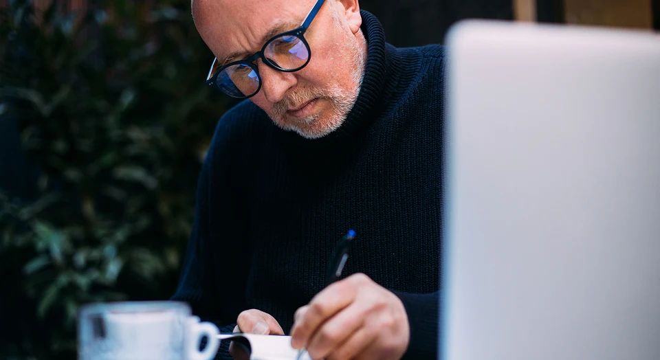 Mann mit Glatze und schwarzer Brille arbeitet am Laptop und schreibt parallel etwas auf Papier