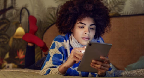Lockige Frau liegt auf ihrem Bauch in ihrem Bett und schaut in ihr Tablet