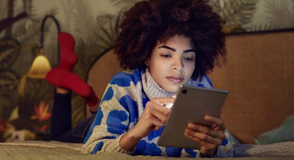 Lockige Frau in buntem Pullover liegt auf dem Bauch auf ihrem Bett und schaut in ihr Tablet
