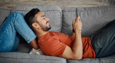 Mann im orangenen T-Shirt liegt auf der Couch mit angewinkelten Beinen und hält ein Smartphone in den Händen
