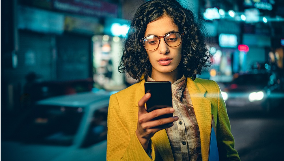 Dunkelhaarige Frau in gelbem Blazer steht abends draußen und schaut in ihr Smartphone