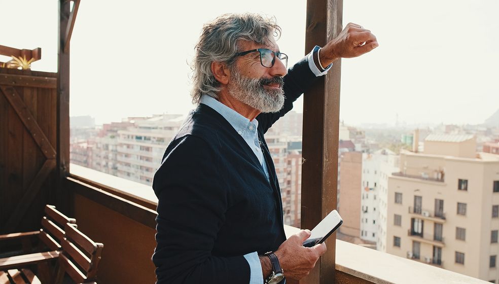 Ein Mann mit grauen Haaren steht auf einem Balkon und blickt über eine Stadtlandschaft. Er trägt ein dunkles Sakko und ein hellblaues Hemd. In der rechten Hand hält er ein Smartphone, mit dem linken Arm lehnt er sich an ein Balkongeländer. Im Hintergrund sind mehrere mehrstöckige Wohngebäude und ein heller Himmel zu sehen. Links im Bild steht eine Holzbank.