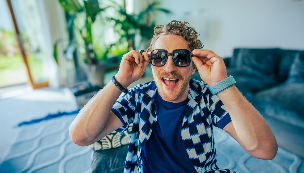 Eine Person sitzt in einem Wohnzimmer mit einem blauen Sofa und grünem Pflanzen im Hintergrund. Die Person trägt ein blau-gemustertes Hemd, ein blaues T-Shirt und ein blaues Armband. Sie hebt beide Hände in Richtung Kopf. Im Hintergrund ist durch ein Fenster Tageslicht zu sehen.