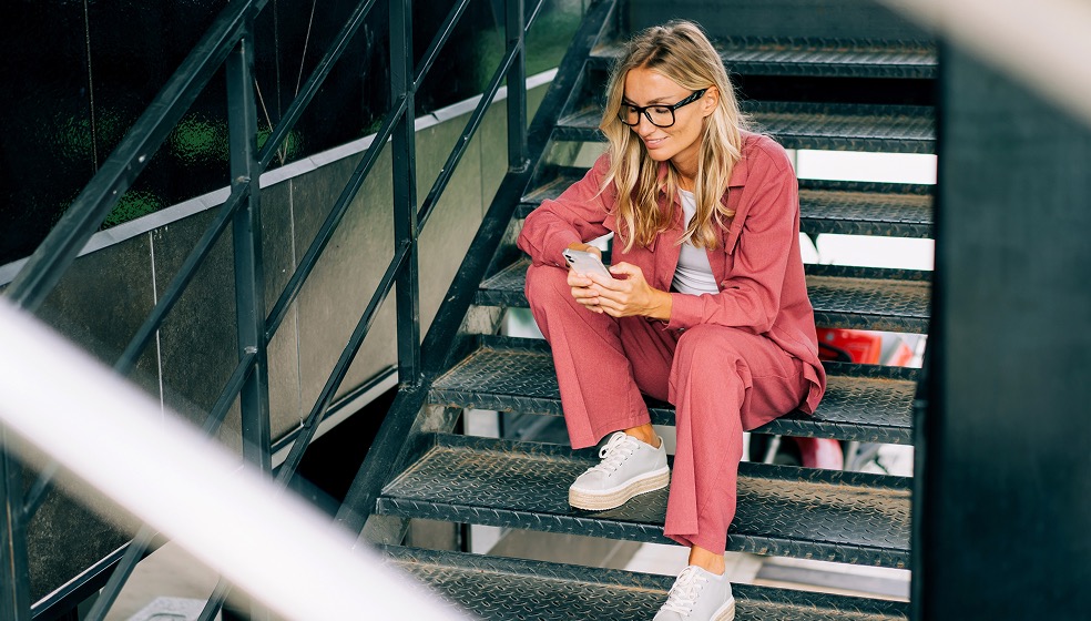 Frau im rosa Anzug mit blonden Haaren und Brille sitzt auf Treppenstufen und blickt nachdenklich lächelnd auf ihr Smartphone, dass sie vor sich in den Händen hält