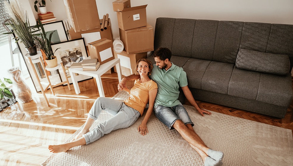 Frau und Mann liegen an Sofa angelehnt in einer frisch eingerichteten Wohnung und lächeln sich an
