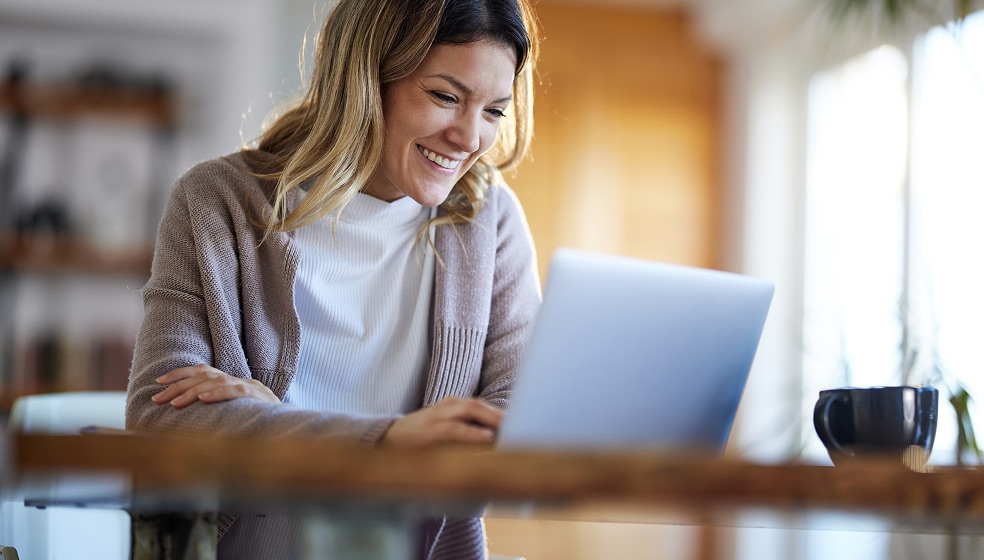 Blonde Frau in Cardigan steht in der Küche lächelt in ihren Laptop