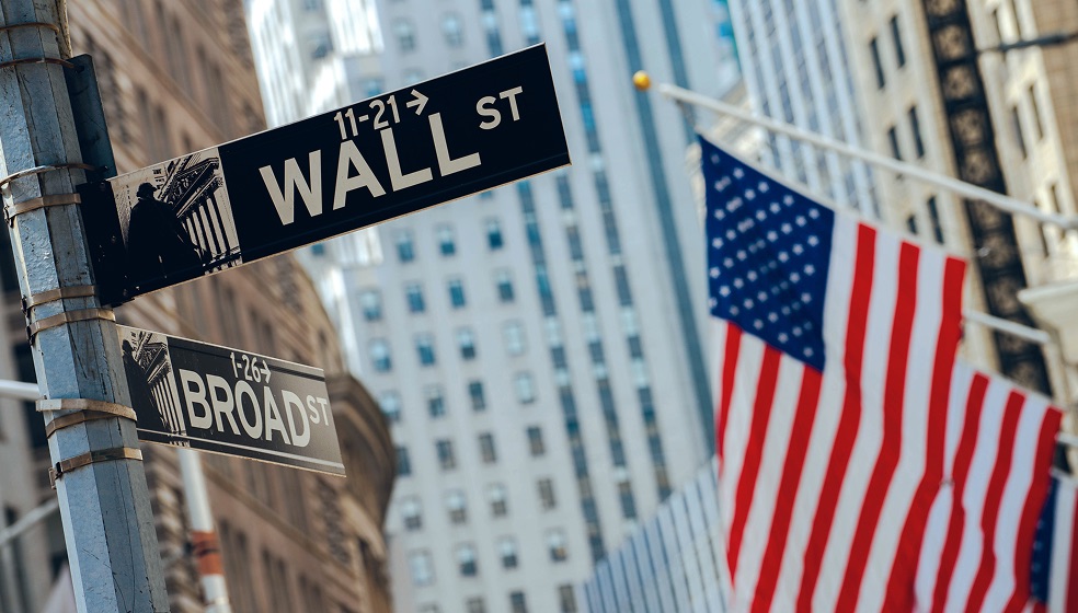 Blick in die Staße der Wallstreet mit Bürogebäuden im Hintergrund. Im Vordergrund sieht man rechts eine amerikanische Flagge, links das Straßenschild der Wallstreet. 