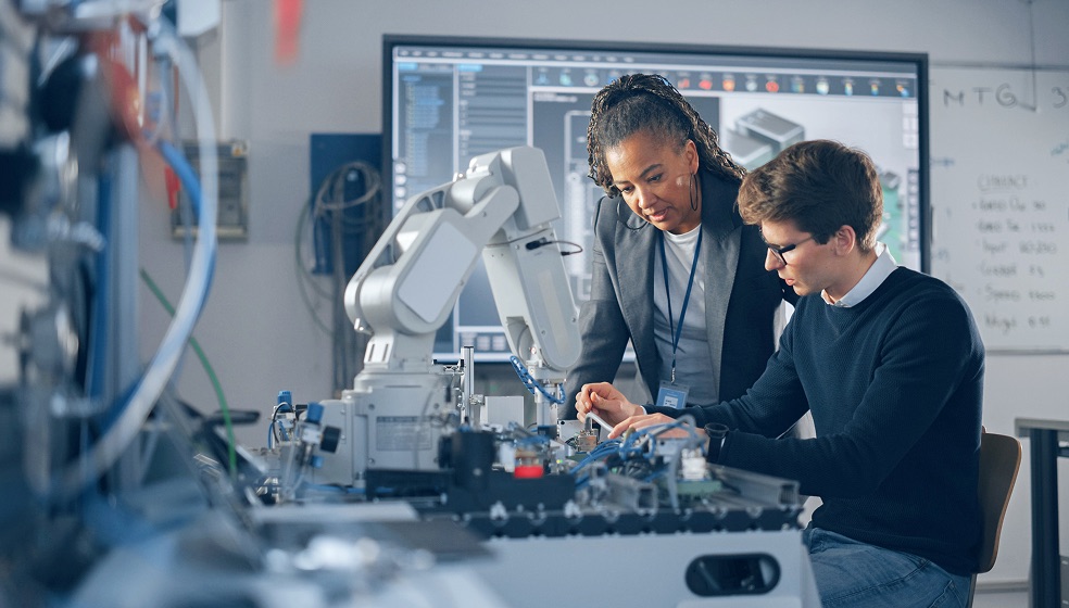 Ein Mann und eine Frau in einem Technologie Labor diskutieren gerade etwas. Vor ihnen befindet sich ein Roboterarm.