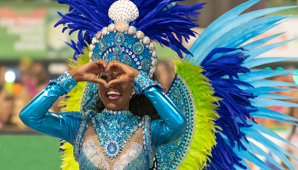 Frau in blau grünem Samba-Outfit formt vor ihrem Gesicht ein Herz mit ihren Händen