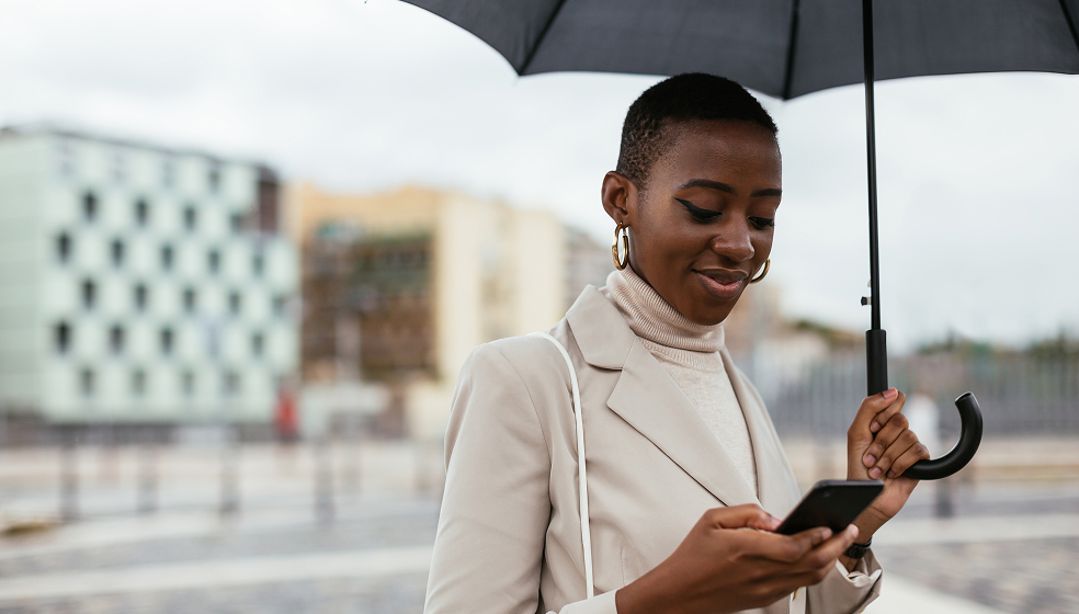 Frau mit Mantel und Regenschirm steht im Freien und schaut auf ihr Smartphone