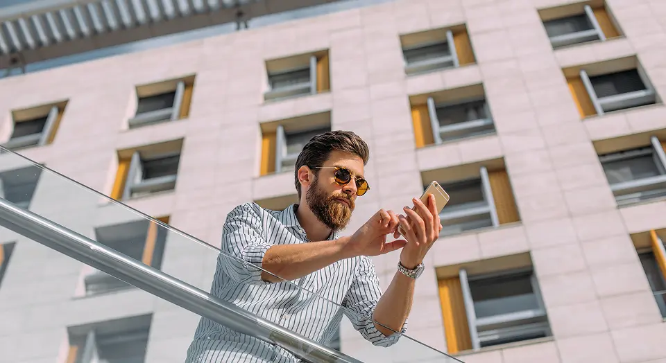 Mann in Hemd mit Sonnenbrille steht draußen an einem Geländer und schaut auf sein Smartphone