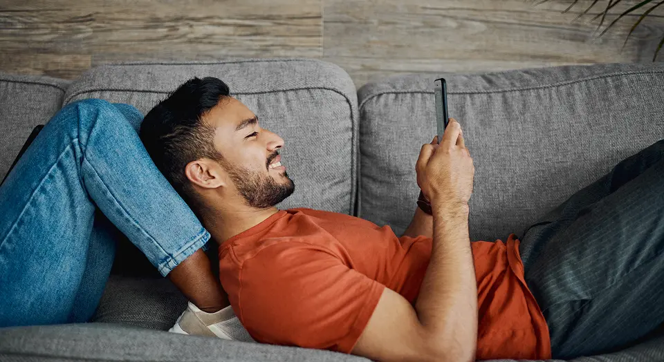 Mann in orangenem T-Shirt liegt auf einem Sofa und schaut in sein Smartphone