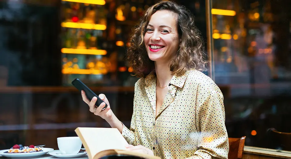 Lockige Frau in Hemd sitzt mit Notizbuch in einem Café und lächelt in die Kamera