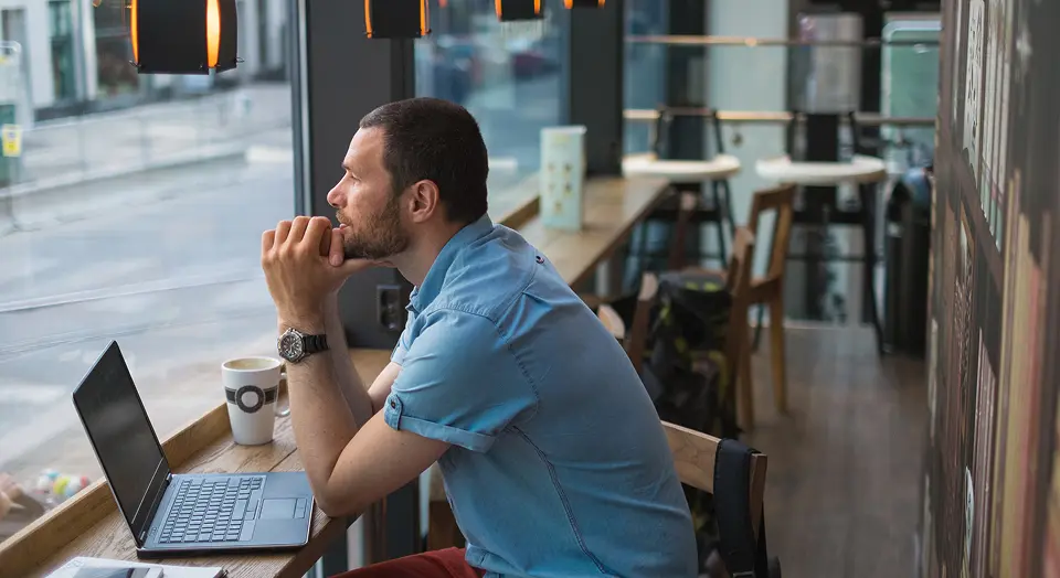 Mann in blauem Hemd sitzt in einem Café am Laptop und aus dem Fenster hinaus