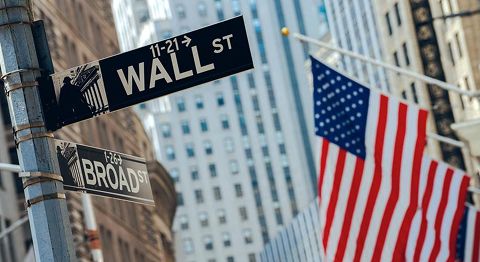 Das Bild zeigt ein Straßenschild mit der Aufschrift „Wall Street“ vor unscharfem Hintergrund mit Hochhäusern und einer amerikanischen Flagge.