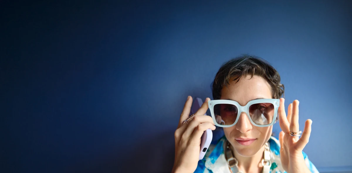Dunkelhaarige Frau mit Sonnenbrille vor einer dunkelblauen Wand mit Smartphone in der Hand
