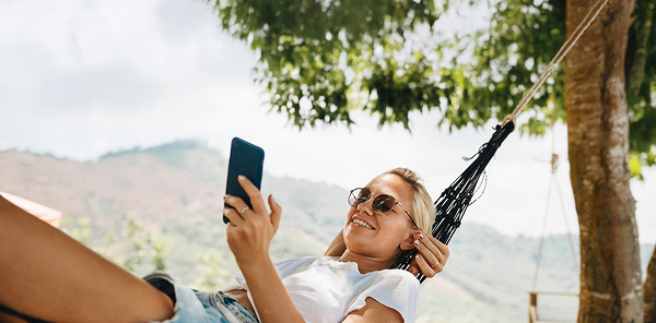Blonde Frau in Sommerklamotten liegt im Freien in einer Hängematte und schaut lächeln in ihr Smartphone