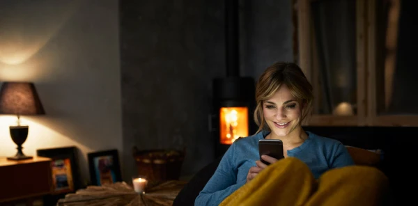 Blonde Frau in blauem Oberteil schaut abends in ihr hell leuchtendes Smartphone und lächelt