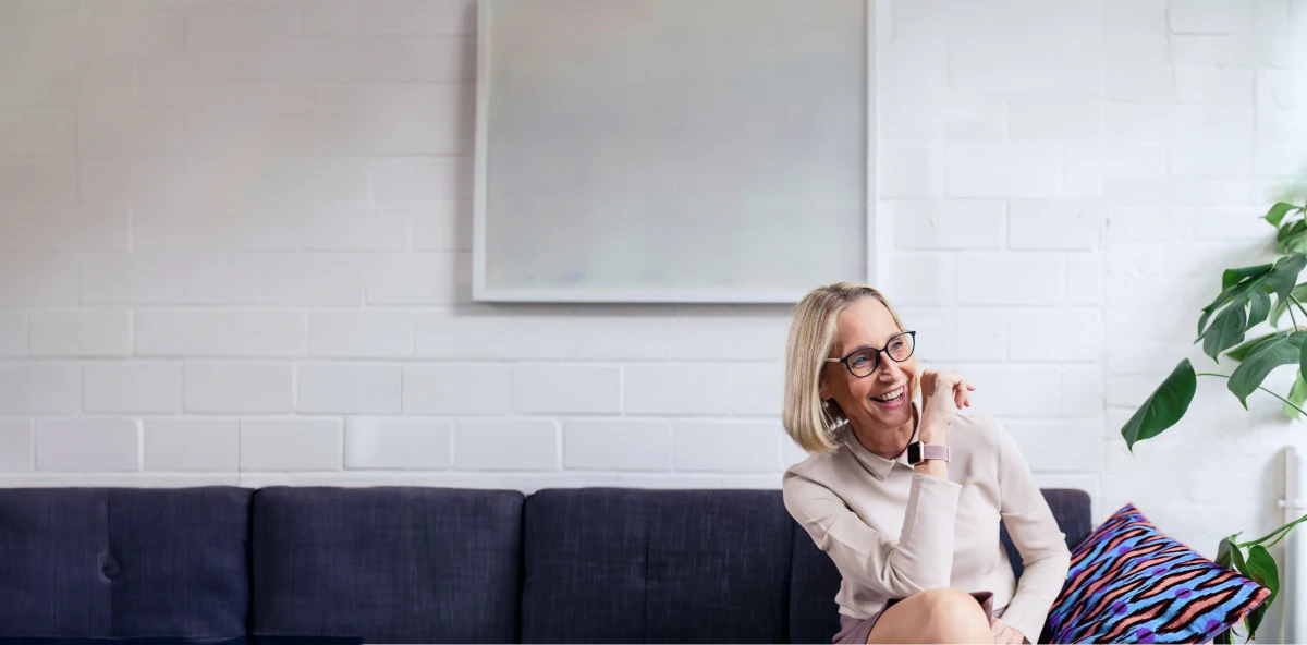 Blonde Frau mit schwarzer Brille sitzt auf beigem Sofa und lacht