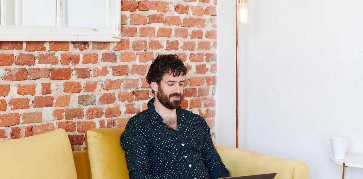 Mann mit dunklen Haaren in schwarzem Hemd mit weißen Punkt sitzt auf einem beigen Sofa und arbeitet am Laptop auf seinem Schoß