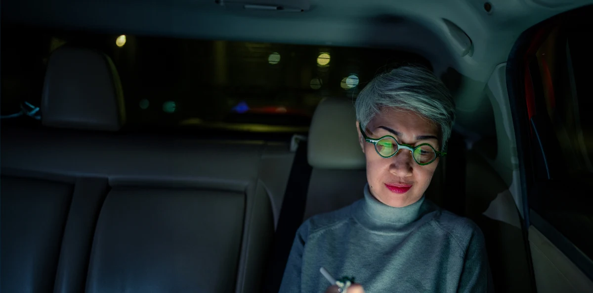 Grauhaarige Frau sitzt abends hinten in einem Auto und tipp auf ihrem Tablet, das sich in ihren Brillengläsern spiegelt